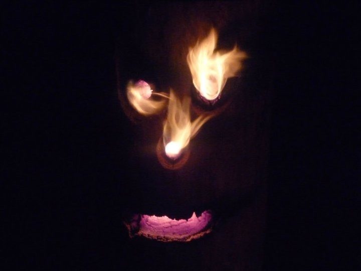 Visage taillé dans une bûche qui brûle la nuit : des flammes sortent des yeux et de la bouche.