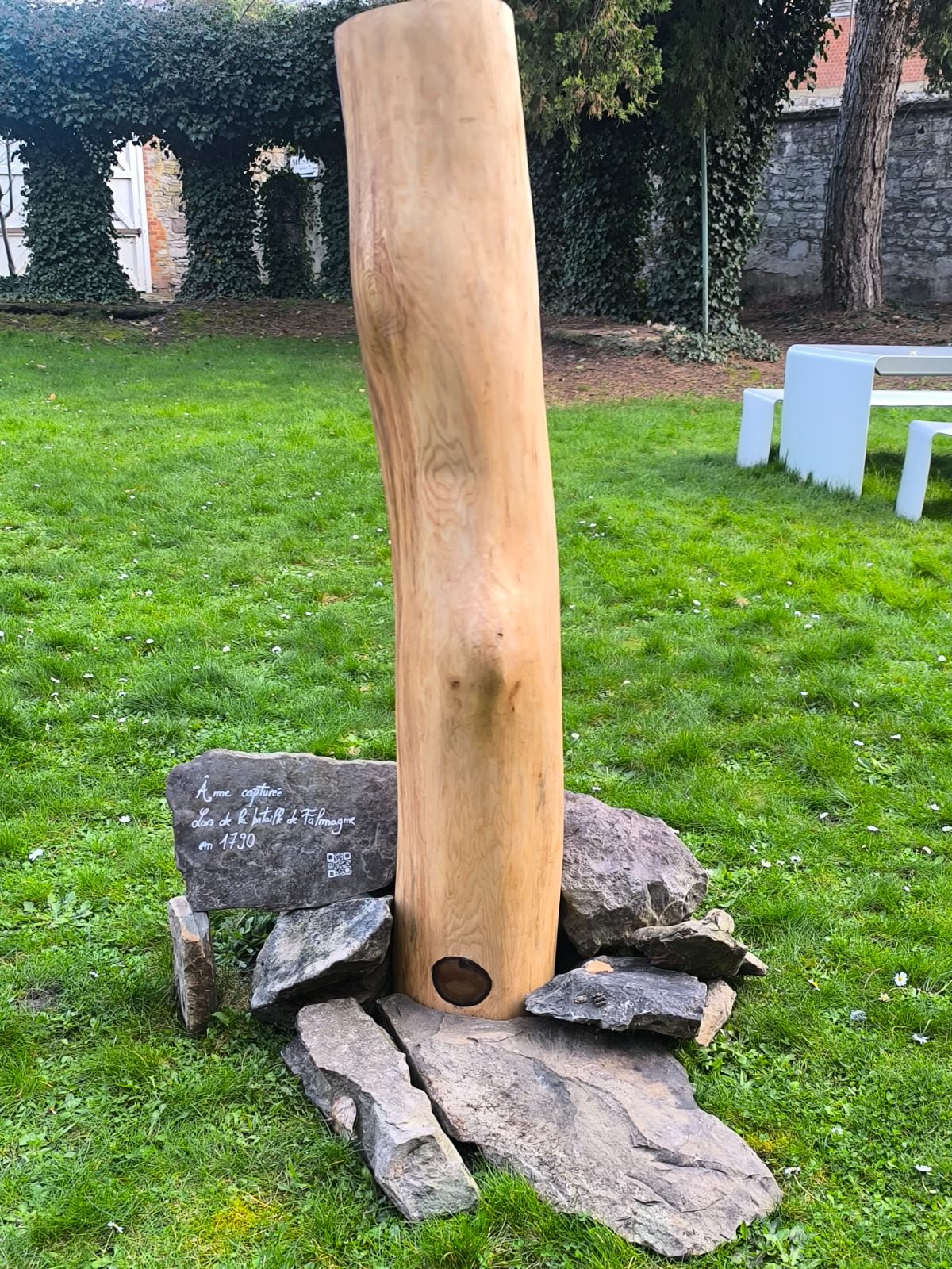 Tronc clair dressé sur des pierres dans un jardin, avec une ardoise gravée « Âme capturée lors de la bataille de Falmagne en 1790 ».